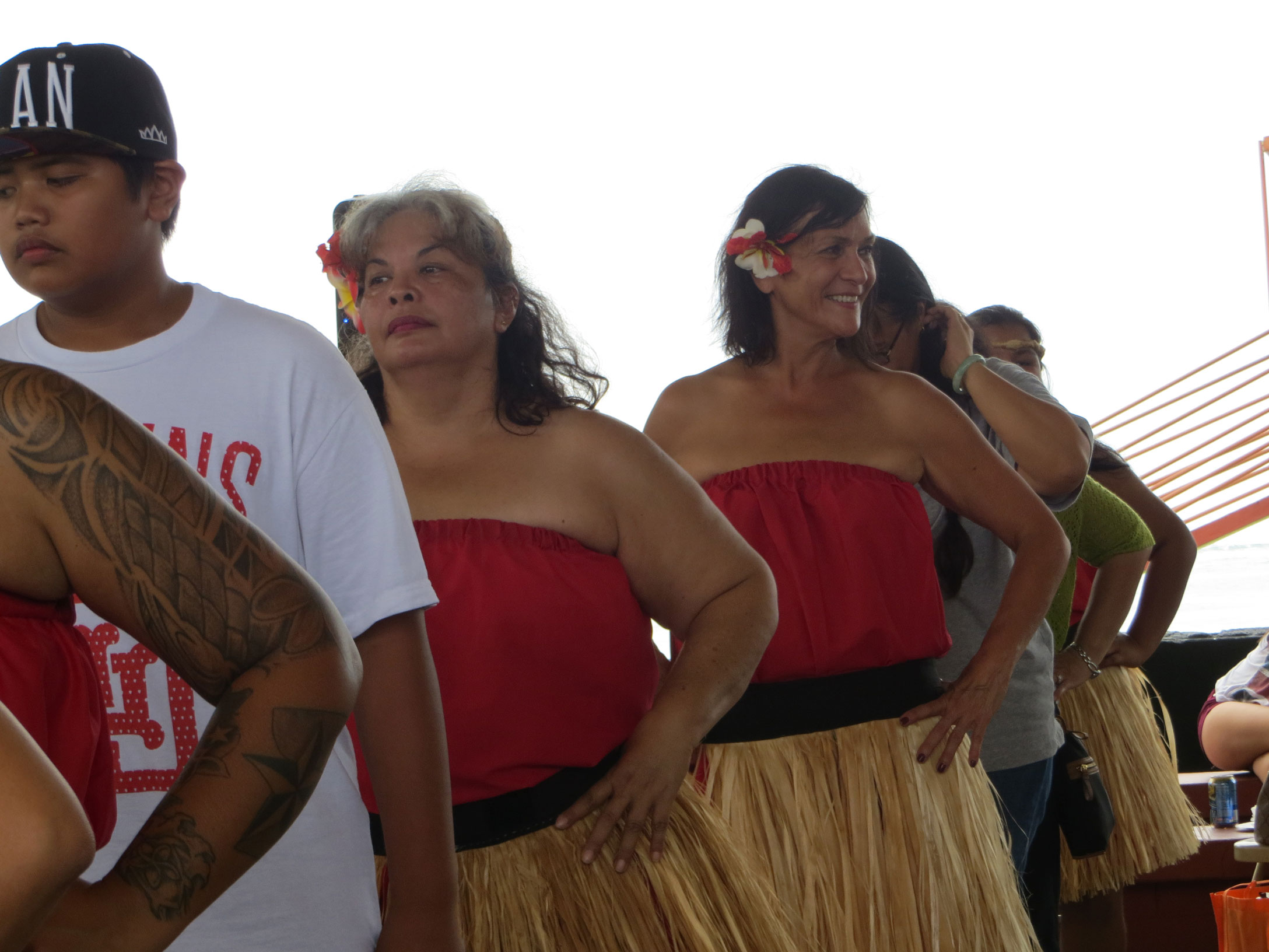 Several individuals standing in single file line wearing grass skirts, red tube tops and a flower in their right ear.